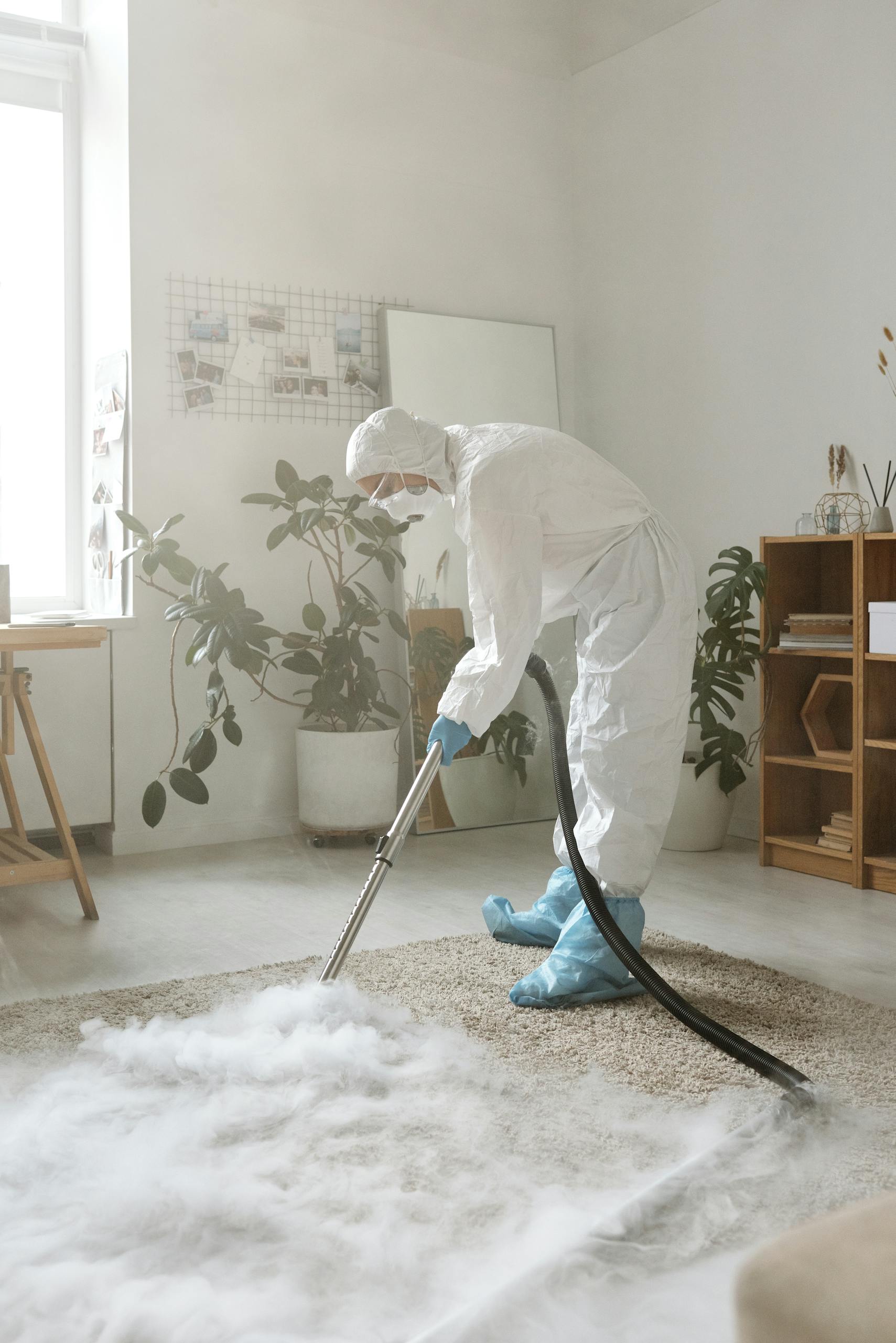 A person in full protective gear disinfects a carpet in a bright indoor space.